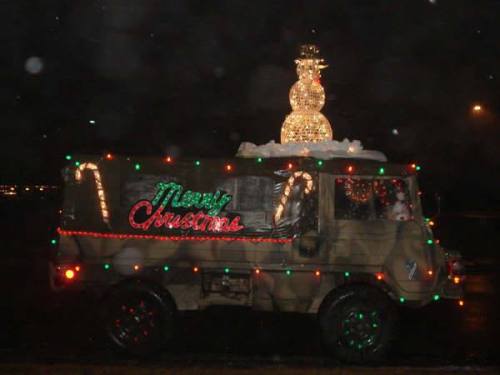 A decorated military truck with Christmas lights and a snowman on top, driving at night.