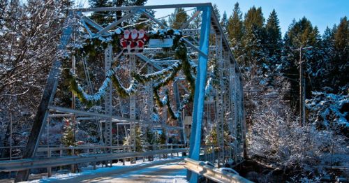 A snowy bridge decorated with Christmas wreaths and ornaments, surrounded by trees and a clear blue sky.