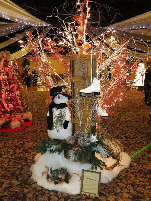 A festive display featuring a snowman, decorated branches, and an ice skate, surrounded by white fluff and holiday decor.