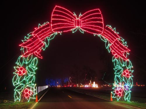 A colorful archway of red and green lights shaped like a bow and holly, welcoming visitors along a dark road.