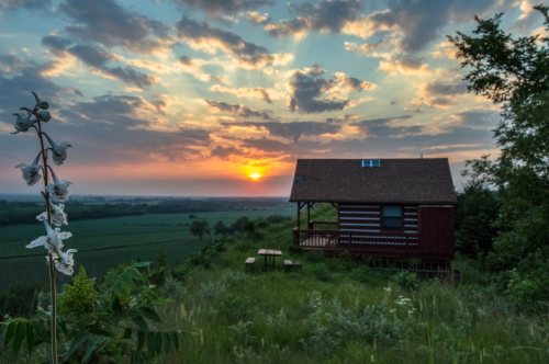A rustic cabin on a hill at sunset, surrounded by lush greenery and a colorful sky filled with clouds.
