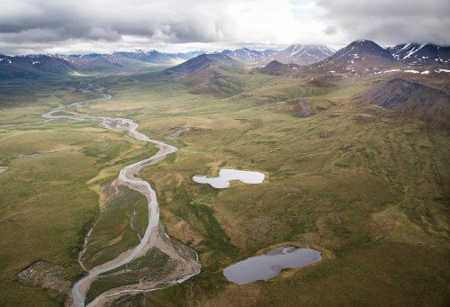 Aerial view of a winding river through a vast, green landscape with mountains and scattered ponds under a cloudy sky.