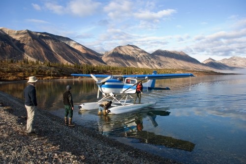 A seaplane on calm water near a rocky shore, with two people working on the plane and another observing nearby.
