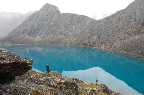Two hikers stand by a stunning turquoise lake surrounded by rugged mountains and misty clouds.