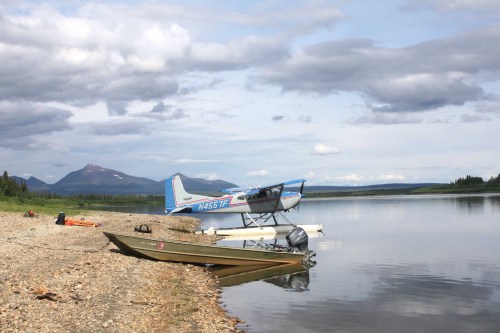 A seaplane parked by a calm river, with a wooden boat on the shore and mountains in the background under a cloudy sky.