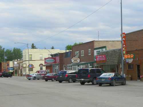 A small town street lined with shops and restaurants, featuring parked cars and a cloudy sky.