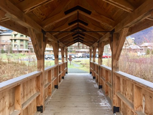 A wooden walkway leads through a covered bridge, surrounded by greenery and buildings in the background.