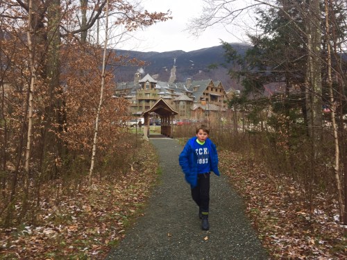 A boy in a blue hoodie runs along a gravel path surrounded by trees, with a large building and mountains in the background.