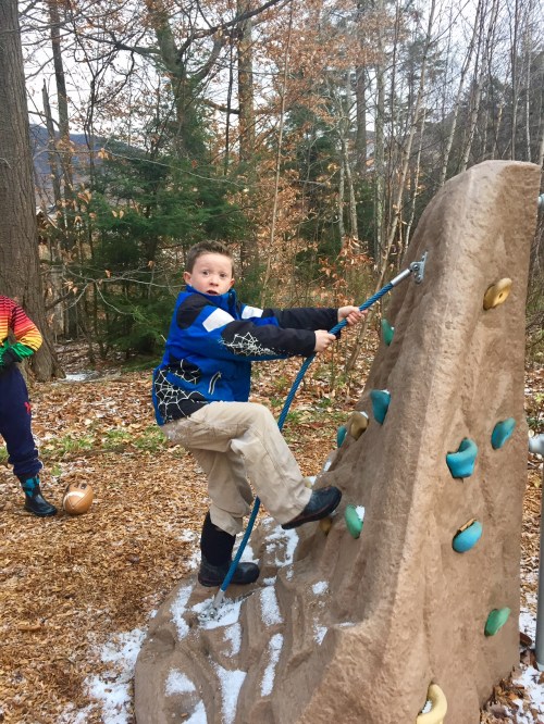 A child climbs a rock wall in a forested area, wearing a blue jacket and beige pants, with fallen leaves around.