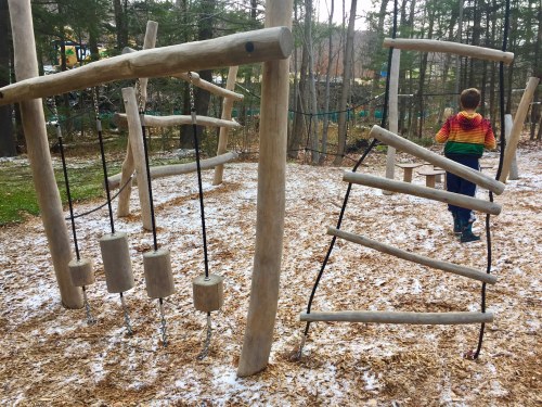 A child in a colorful sweater walks past a wooden playground structure with swings and climbing elements, surrounded by snow.