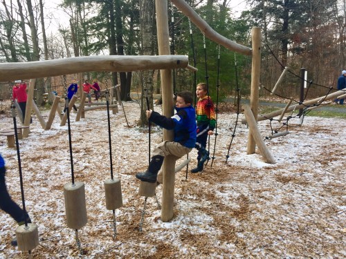 Children play on a wooden playground structure in a snowy, wooded area, enjoying swings and climbing activities.