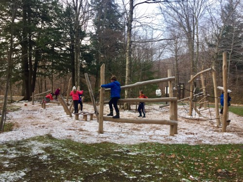 Children playing on a wooden playground structure in a snowy forested area. Trees surround the play area.