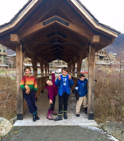 Four children playfully posing under a wooden structure in a scenic outdoor setting.