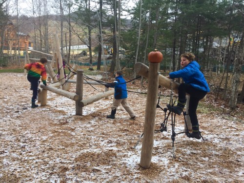 Children play on a wooden climbing structure in a forested area, with some snow on the ground and trees in the background.