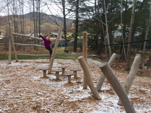 A child plays on a wooden playground structure in a forested area with fallen leaves and a snowy ground.
