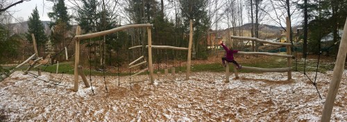 A child in a purple jacket plays on a wooden climbing structure in a snowy, wooded area.