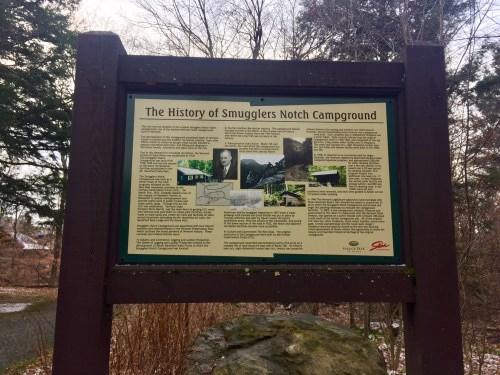 Sign detailing the history of Smugglers Notch Campground, surrounded by trees and a gravel path.