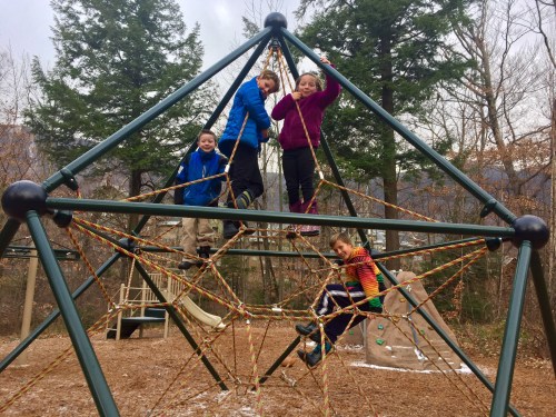 Four children play on a climbing structure in a park, surrounded by trees and a slide in the background.