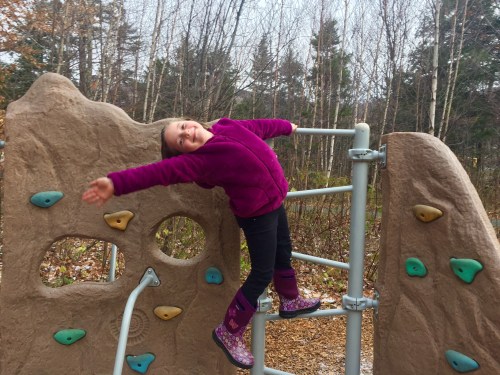 A young girl in a purple jacket joyfully hangs from a playground climbing structure in a wooded area.