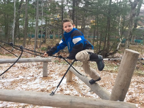 A young boy in a blue jacket climbs on a wooden structure in a forested area with fallen leaves and snow.