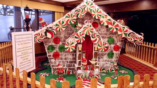 A festive gingerbread house decorated with candy, surrounded by a picket fence and a sign about the display.