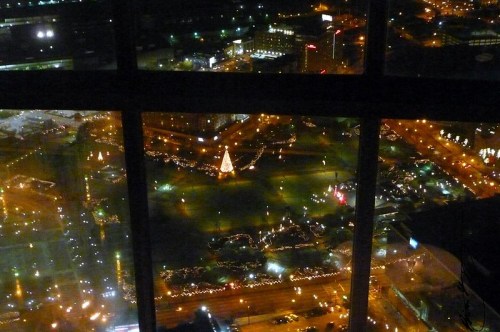 Aerial view of a city park decorated with holiday lights, featuring a Christmas tree, seen through a window at night.