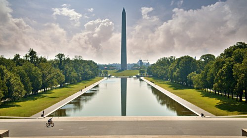 A view of the Washington Monument reflecting in the pool, surrounded by trees and a clear sky. A cyclist rides nearby.