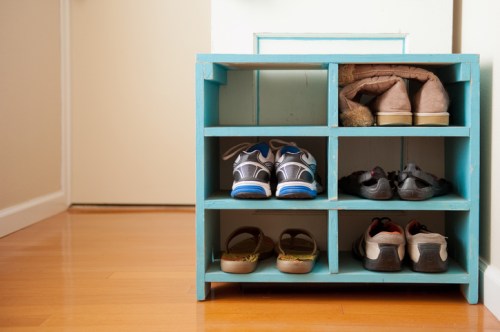 A blue shoe rack with six compartments, holding various pairs of shoes, on a wooden floor near a doorway.