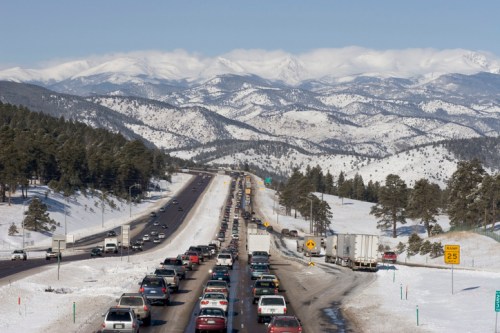 Traffic jam on a snowy highway with mountains in the background, surrounded by pine trees and a clear blue sky.