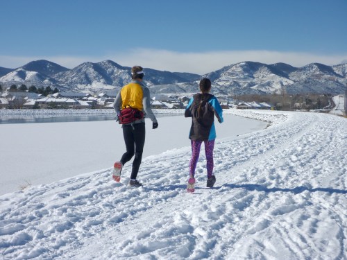 Two runners jog along a snowy path beside a frozen lake, with mountains in the background under a clear blue sky.