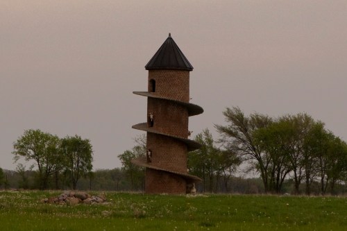 A unique spiral brick tower stands in a grassy field, surrounded by trees under a cloudy sky.