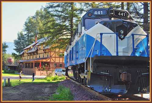 A blue and white train engine next to a rustic building, with trees in the background and people walking nearby.