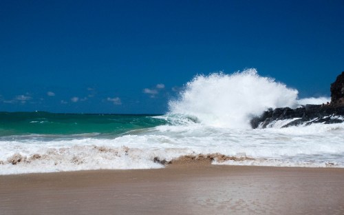 Waves crashing against a rocky shore under a clear blue sky, with sandy beach in the foreground.