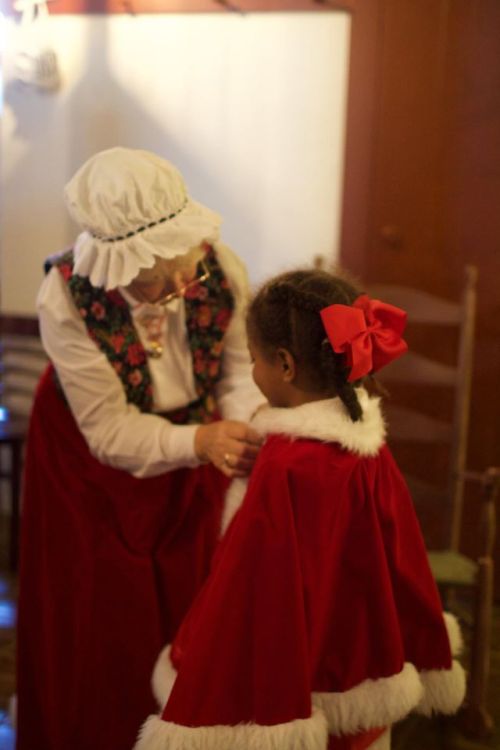 An elderly woman in a festive outfit adjusts a red cape on a young girl with a bow in her hair.