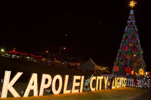 Colorful Christmas tree adorned with lights next to a sign reading "Kapolei City Lights" at night.