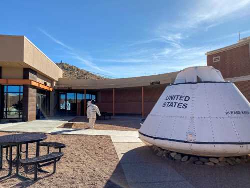 An astronaut in a spacesuit walks past a spacecraft display outside a museum in a desert setting.