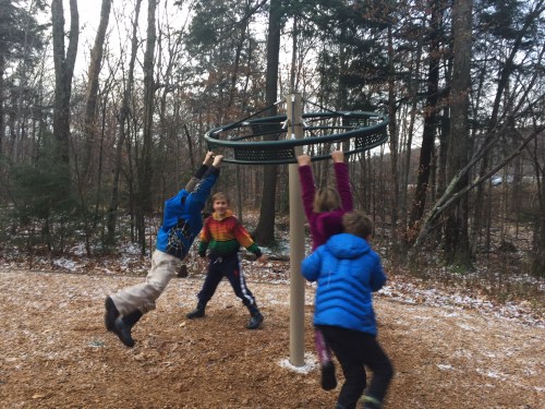 Four children play on a spinning playground structure in a wooded area, with some snow on the ground.