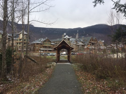 A pathway leads to a wooden archway, with a mountain resort and trees in the background under a cloudy sky.