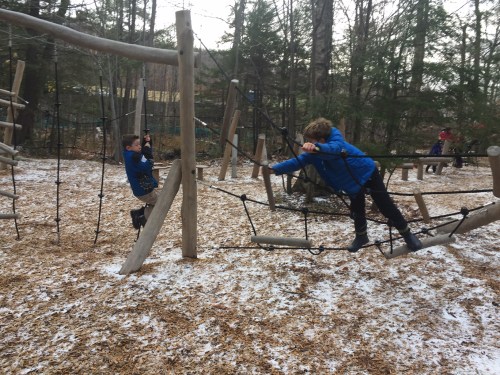 Two children play on a wooden climbing structure in a snowy playground surrounded by trees.