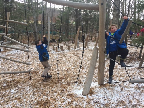 Two children play on a wooden climbing structure in a forested area, surrounded by fallen leaves and snow.