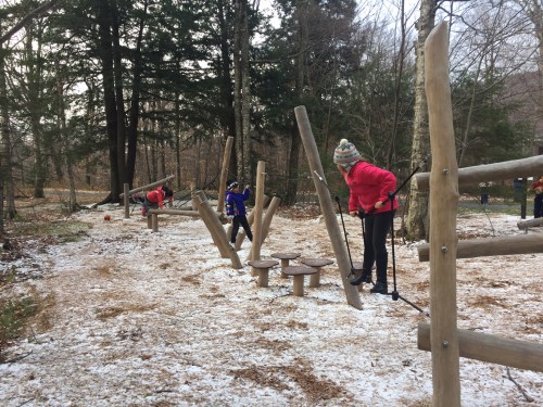 Children play on a wooden obstacle course in a snowy forest, climbing and balancing on various structures.