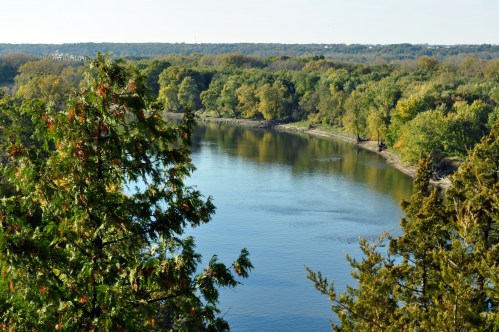 A serene river flows through a lush green landscape, surrounded by trees under a clear blue sky.