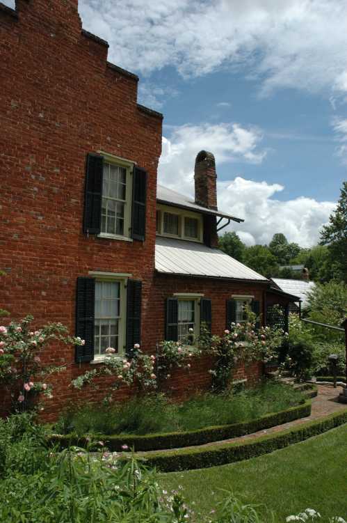 A charming red brick house with green shutters, surrounded by blooming roses and lush greenery under a blue sky.