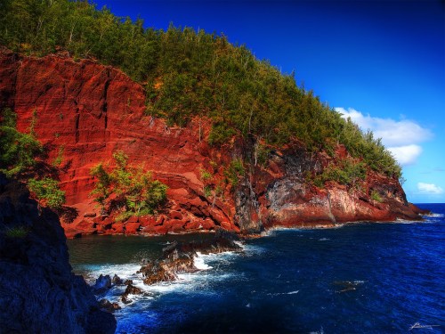 Vibrant red cliffs rise above a blue ocean, with lush greenery along the shoreline under a clear sky.