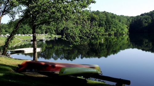 Two canoes rest on the grass by a calm lake, surrounded by trees and reflecting the serene landscape.