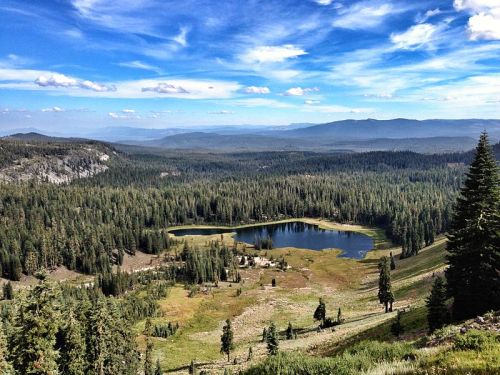 A scenic view of a lush green valley with a serene lake, surrounded by trees and mountains under a blue sky.