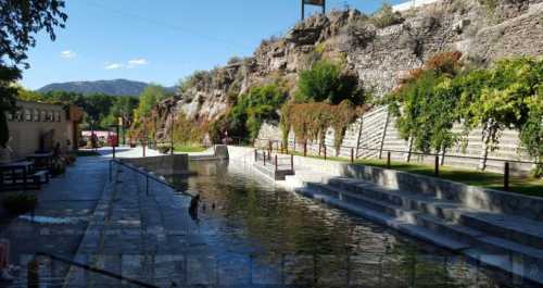 A serene landscape featuring a stone pathway alongside a calm waterway, surrounded by greenery and rocky hills.