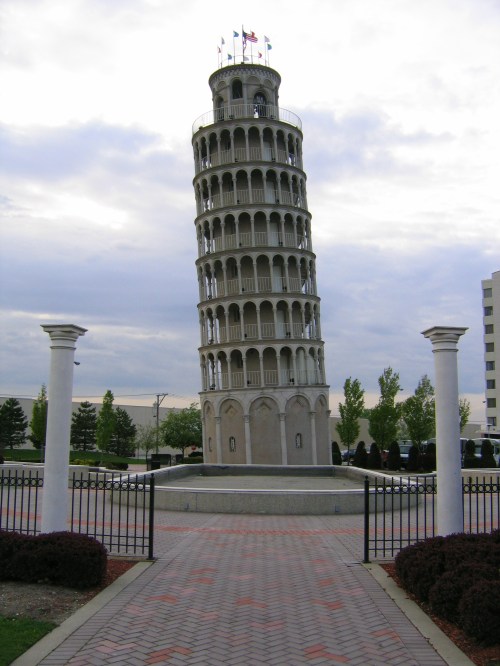 A replica of the Leaning Tower of Pisa stands in a park, surrounded by greenery and a pathway leading to it.
