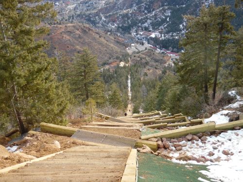 A steep, wooden staircase descends through a forested area, leading down a mountain with a view of a valley below.