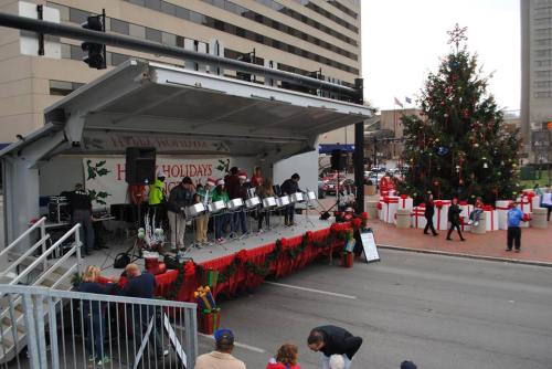 A holiday event stage with musicians playing instruments, surrounded by festive decorations and a Christmas tree.